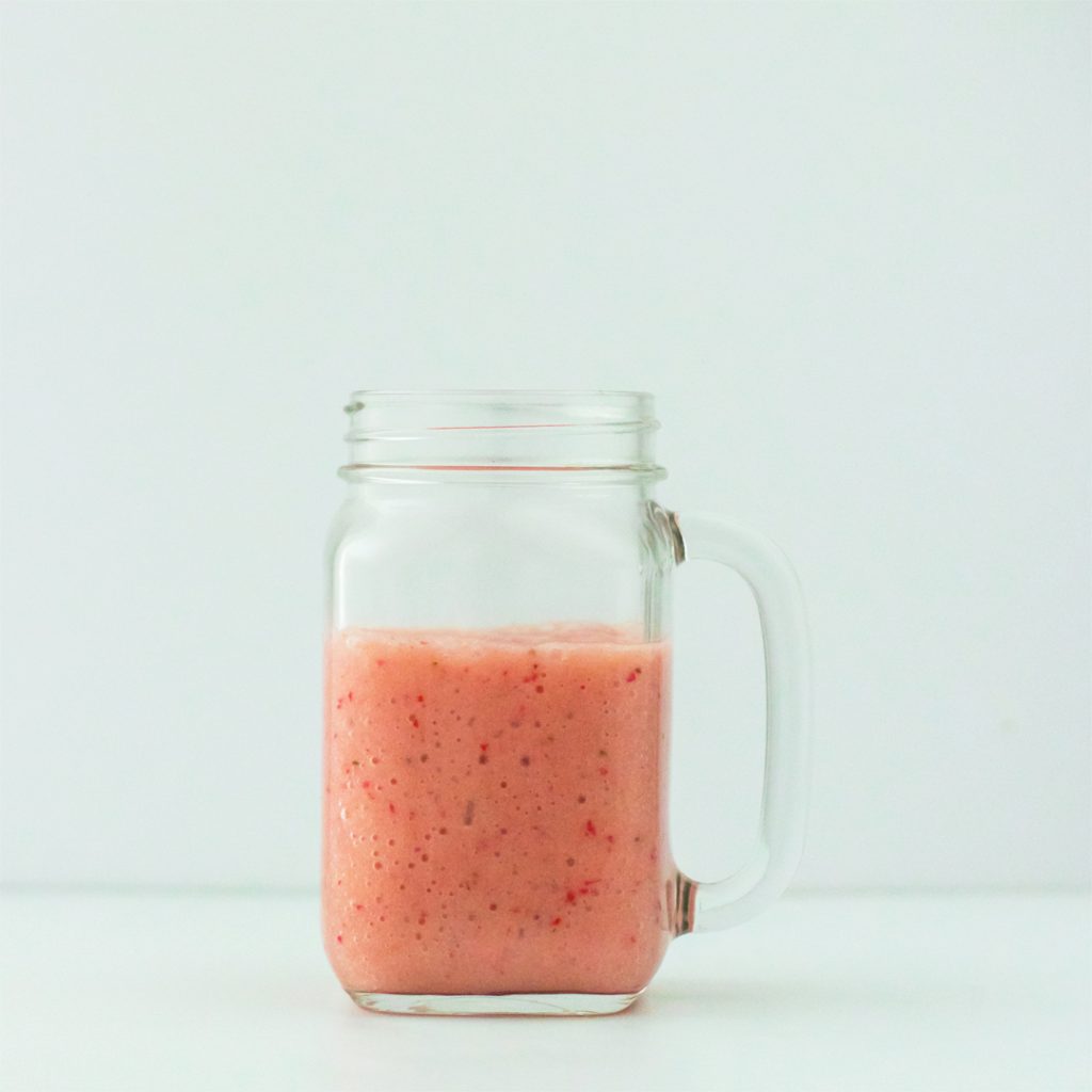 Vaso transparente sobre mesa blanca. Fondo blando. En el vaso bebida rosada con motas rojizas de las fresas.