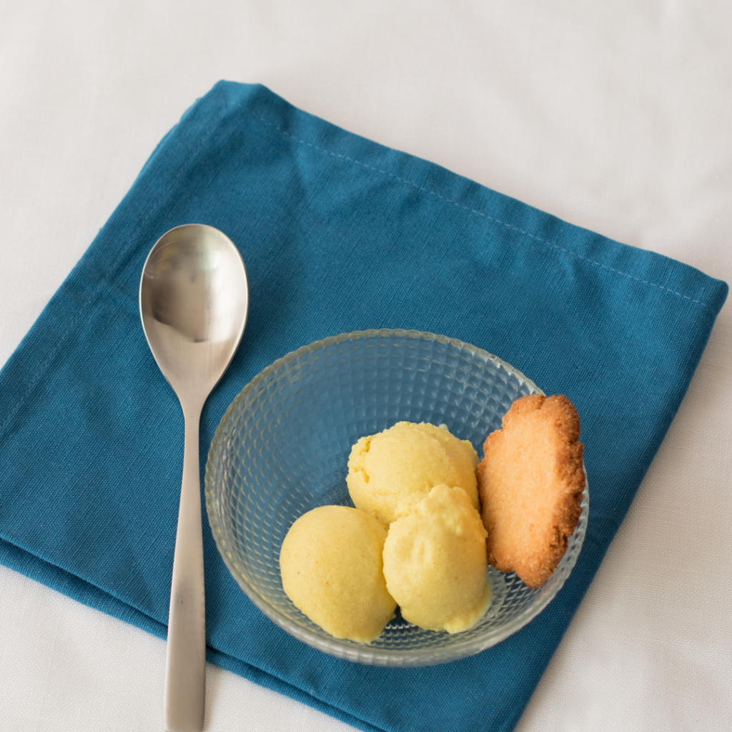 Tres bolas de helado cremosos de leche dorada acompañadas de una galleta. Cuchara plata. Todo sobre servilleta azul mar, en mesa blanca.